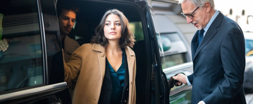 chauffeur holding the car door for the lady to get down. A co passenger is also following her.