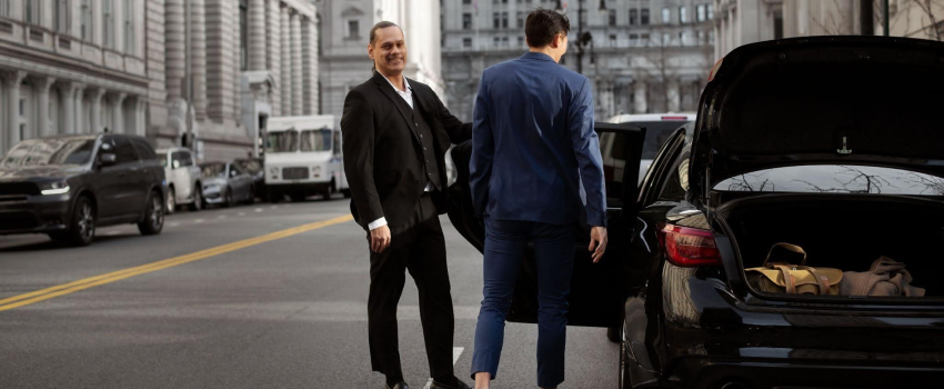 A chauffeur is welcoming an executive passenger with a warm smile and opening the car door for him.
